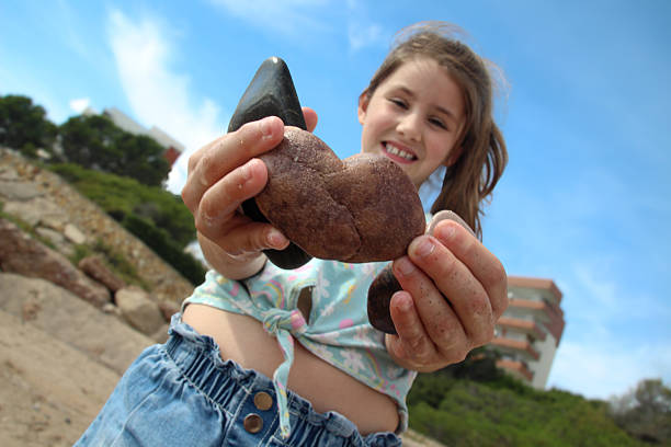A young girl holding a large shell at the beach, smiling, with a blue sky and buildings in the background