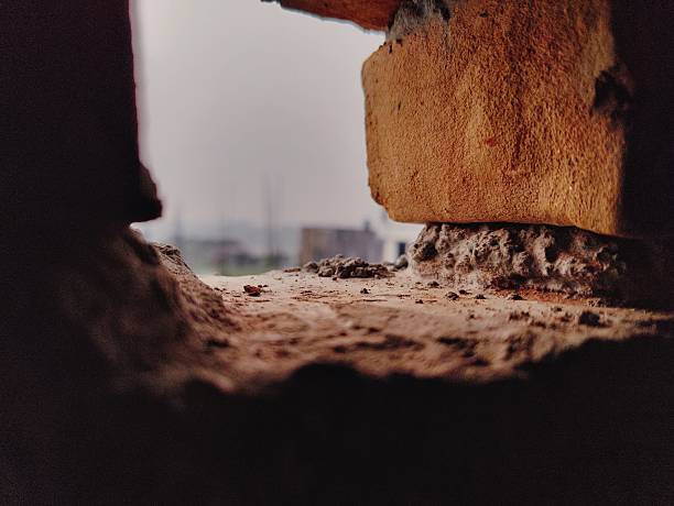 A gritty close-up through a brick opening reveals dusty ground and rubble at a construction site, with a distant urban backdrop. Raw texture, weathered materials, and industrial atmosphere dominate the scene.