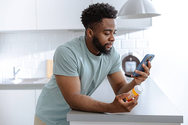istock Young man at the kitchen counter reads medication bottle while holding a smartphone. 2253525884