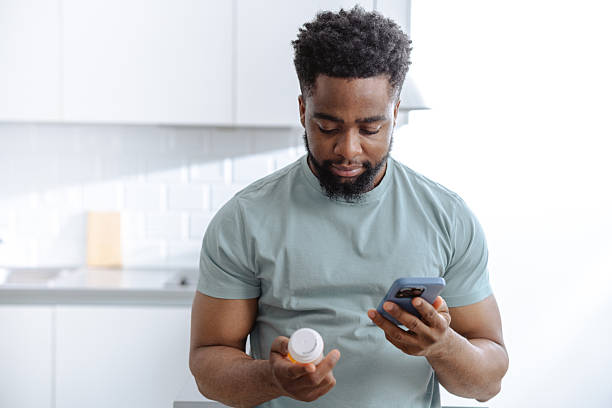 istock Young man in a modern kitchen reviews medicine on his phone while holding a pill bottle 2253525895
