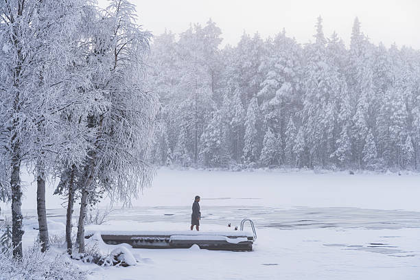 Brave swimmer wrapped in bathrobe walks in the snow on a pier above the frozen water of a mountain lake in winter, Finnish Lapland, Finland