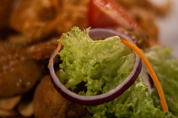 Close-up of a fresh garnish including leafy lettuce, red onion ring, tomato and fried meat in the background. The image blends textures and colors in a visually rich food presentation.