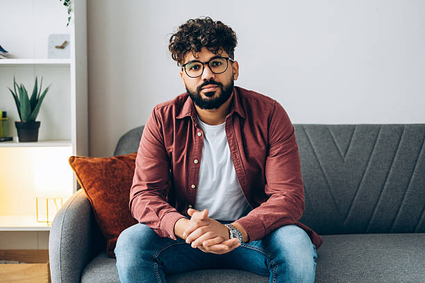 istock Young handsome serious man with glasses sitting on couch at home looking at camera 2253531122