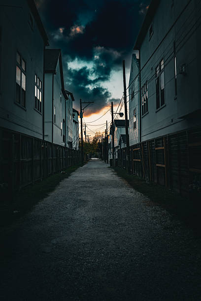 A cinematic, low-angle perspective of a narrow gravel alleyway lined with modern white residential buildings and wooden fences.