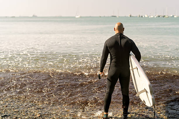 Bald man in wetsuit holding a surfboard at the shoreline, facing the ocean and ready for surfing on a sunny beach day