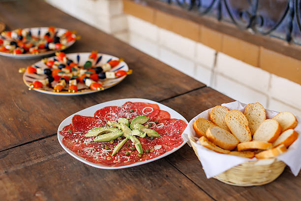 Mediterranean appetizer table with beef carpaccio topped with avocado and capers, served with sliced bread and antipasti plates on a rustic wooden table.