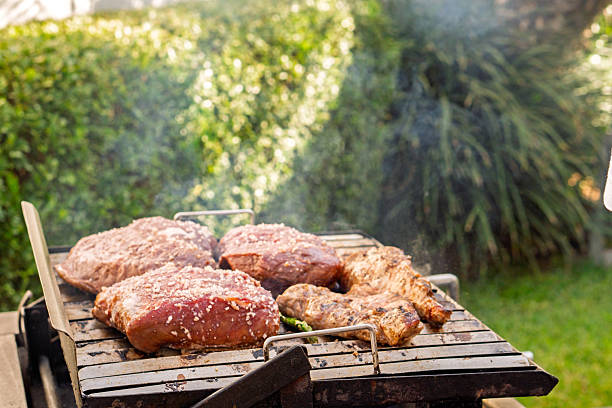 Three large cuts of beef and two pieces of chicken cooking on a hot grill, generating smoke in a backyard garden