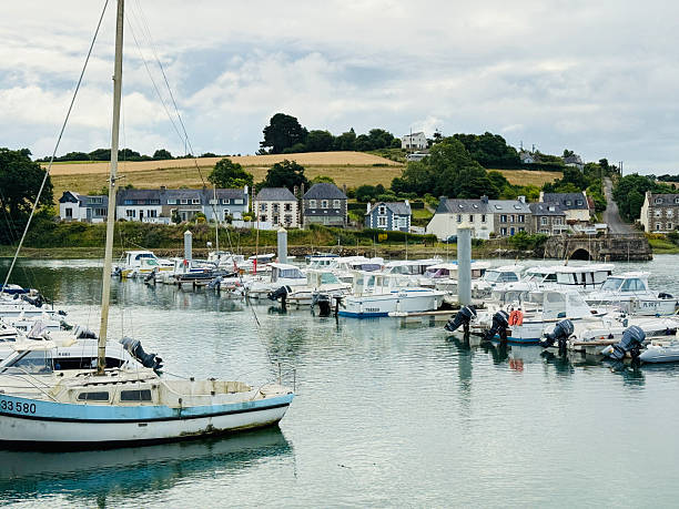 Horizontal photo of sailing boats, reflecting on the calm waters of the Jaudy River, moored at the floating pontoons in the deep water harbour, at the historic town of Treguier on the Pink Granite Coast in Brittany. Traditional stone houses at the water's edge in the background.