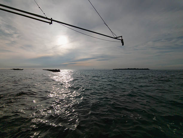 A peaceful ocean scene with sunlight reflecting on gentle sea waves, distant boats on the horizon, and an overhead fishing structure framing the sky. The image captures a calm maritime atmosphere with soft clouds and a quiet coastal environment.