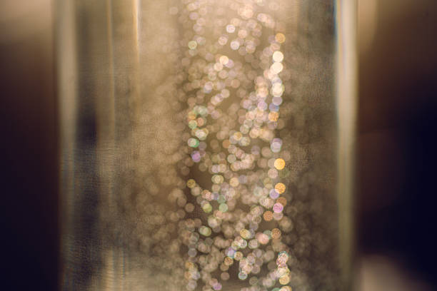 Many small, shimmering bubbles rising & clinging to the inside of glass filled with carbonated beverage, creating abstract bokeh, illuminated by soft sunlight casting textured light. Party concept