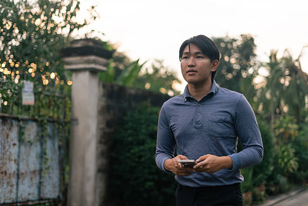 Young asian man holding a smartphone while walking outdoors, looking forward, thinking, traveling
