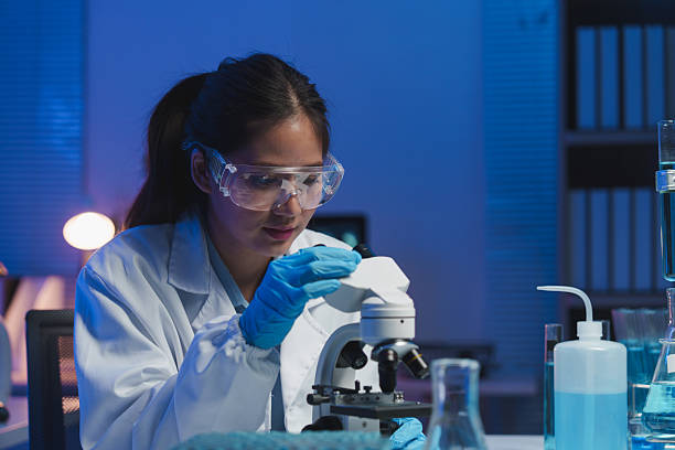 istock Young female scientist adjusting a microscope in a dimly lit laboratory filled with scientific equipment and blue-toned solutions, focused on her research 2253558976