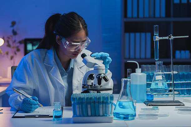 istock Young female scientist wearing lab coat and protective glasses looking through microscope and taking notes during experiment in modern laboratory 2253558983