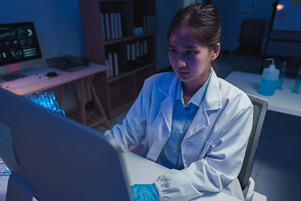 istock Young female scientist wearing a lab coat and gloves is working on a computer in a modern laboratory at night, illuminated by blue light, focused on her research 2253559076