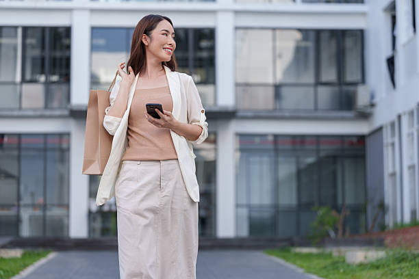 istock Young asian woman walking through the city, smiling while using a mobile phone application and carrying a shopping bag, enjoying a vibrant urban lifestyle on a sunny day 2253560201