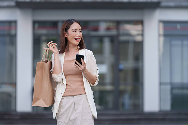 istock Happy customer using a smartphone while holding a shopping bag, looking away and smiling joyfully. Enjoying a moment of satisfaction in an urban setting, radiating positive energy 2253560204