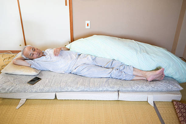 A man is laying on a bed with a pillow and a cell phone on his lap. The bed is a futon, and the man is wearing gray pants. Scene is relaxed and comfortable