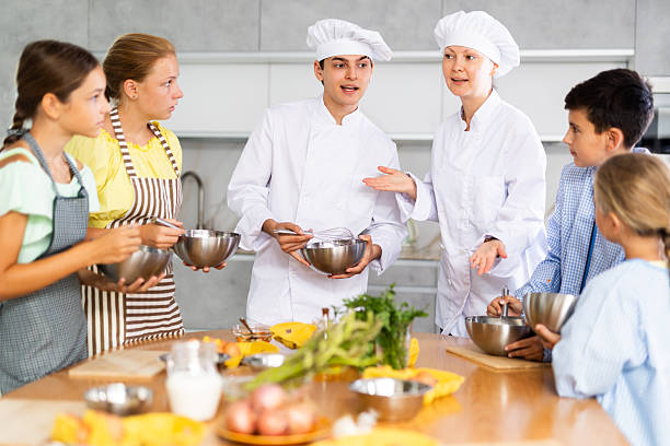 Two experienced chefs, friendly woman and man in white uniform giving culinary lesson to group of inquisitive tweens, teaching basic cooking skills