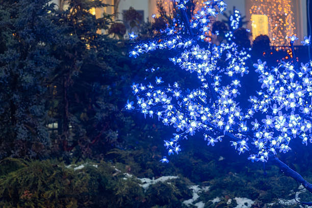 Tree decorated with blue LED flower lights at night. Festive illuminated tree with glowing blue floral lights and warm background in urban celebration setting
