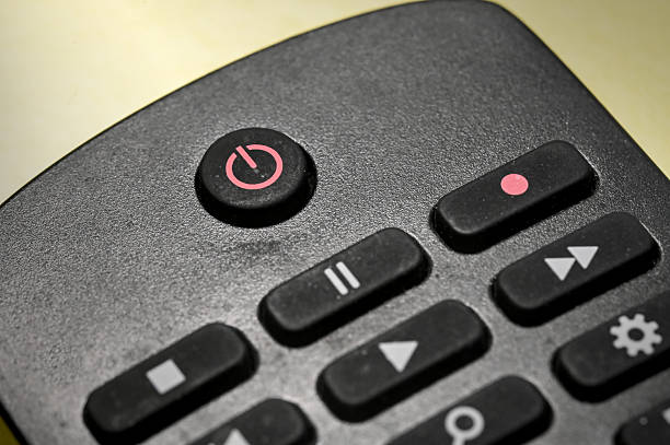 Front view close-up of an unrecognisable young man holding a TV remote control in front of a chroma key studio background. He is pressing buttons on the remote control.