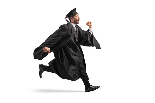 Female student jumps in celebration while wearing a graduation gown and holding a diploma. Vertical shot. Isolated on white.