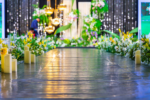 A close-up of clear tall candle holders and lush floral arrangements in yellow and white that run along the table. In the blurred background are round banquet tables with white chair covers and gift bags, warm lighting producing a soft glow. The shallow depth of field emphasizes the texture and colors of the centerpiece while conveying an elegant, festive mood.
