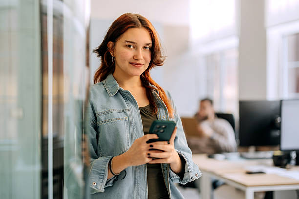 istock Young casually clothed businesswoman posing for camera while using smart phone, leaning on glass wall at the office 2253578305