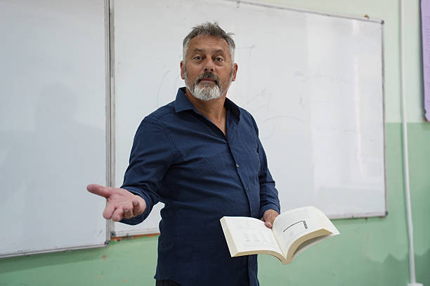 A mature male professor giving a lecture in a classroom, standing in front of a whiteboard while holding a book and gesturing with his hands. He appears engaged and confident while explaining educational content. Concept of education, teaching, academic learning, knowledge transfer, classroom instruction, and professional training.