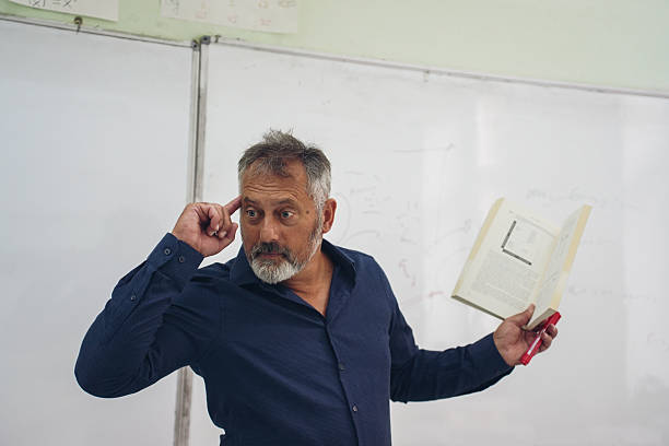 A mature male professor giving a lecture in a classroom, standing in front of a whiteboard while holding a book and gesturing with his hands. He appears engaged and confident while explaining educational content. Concept of education, teaching, academic learning, knowledge transfer, classroom instruction, and professional training.