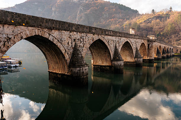 An old stone bridge stretches across the calm Drina River in Višegrad, with its historic arches reflected in the still water.
