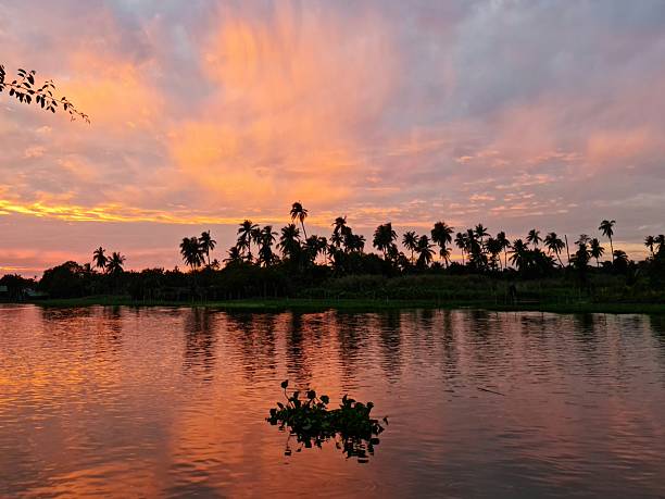 Scenic view of a warm sunset over a calm river in rural Thailand, with soft golden light reflecting on the water and silhouettes of natural landscape.