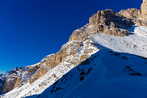 Deserted steep winding ski slope lined with a red protective barrier in the Alps on a sunny winter day. A yellow snow machine is along the run.