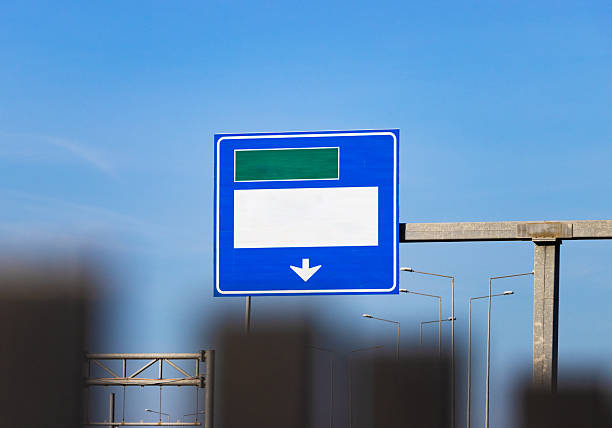 Editable highway road sign with blank green and white panels—mounted on metal structure above streetlights, framed by clear blue sky in a modern urban navigation and infrastructure scene.