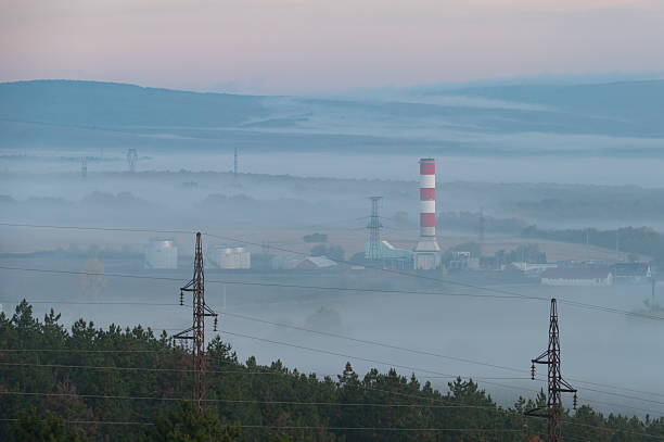 A misty valley landscape captured in early morning light with rolling hills fading into the distance. An industrial power plant with a red and white chimney rises above the fog, blending infrastructure with the surrounding natural environment.