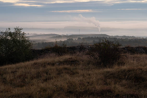 A tranquil rural landscape captured in early morning light with rolling hills covered in mist. In the distance, an industrial chimney releases smoke, creating contrast between nature and human activity.