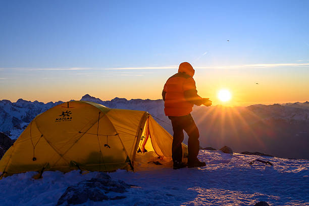 A lone climber beside a yellow tent at sunrise on a snowy mountain peak. The sun casts a warm glow over the snow-covered landscape, highlighting the vast, rugged terrain. The scene captures the solitude and beauty of winter mountain climbing.