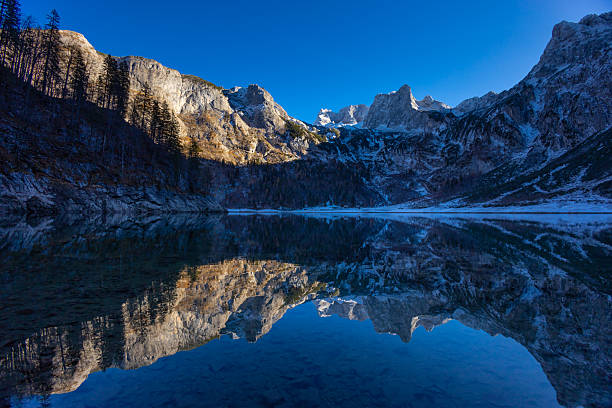 Gosausee lake in Austria with clear alpine water reflecting Dachstein mountains and surrounding forest. Natural mountain landscape showing glacial lake, rocky peaks, outdoor environment, tourism destination, European Alps scenery, nature background, and freshwater ecosystem