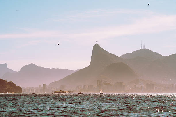 Telephoto seascape of Rio de Janeiro Corcovado, hazy city skyline below, sailboats on sparkling Atlantic water and birds in pastel sky