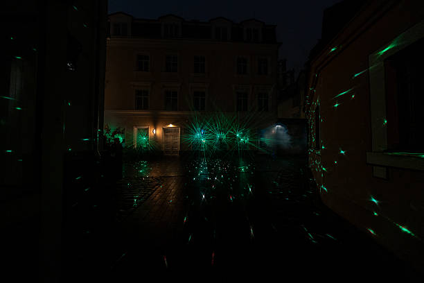 Emerald laser beams burst into star like flares by a three story townhouse, fog intensifies the rays, reflections shimmer on wet cobblestones in low key night light.