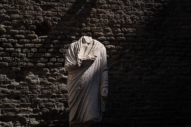 Fragmented marble statue without a head stands against an ancient Roman brick wall, partially illuminated by natural light. The weathered drapery and damaged form emphasize the passage of time, loss, and archaeological history within the ruins of ancient Rome.