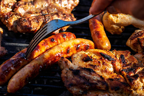 Grilled sausage held with metal tongs over barbecue with assorted meat and vegetable skewers in the background.