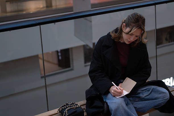 Young woman sitting on a bench inside a modern building and writing in a small notebook with full concentration. Calm lifestyle moment suitable for education topics, journaling habits, creative writing, productivity content, planning and organization themes, remote work visuals, and personal growth materials.