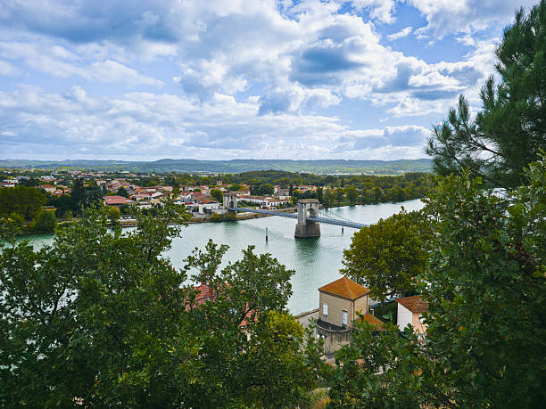 Editorial image showing the historic suspension bridge in Andance, a small town in Ardèche, France. The bridge spans the Rhône river and connects the two riverbanks with a scenic view of the surrounding landscape. This photo was taken on 5 October 2025.