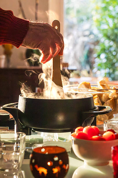 Atmospheric close-up of a pot of steaming cheese fondue on a table. A hand stirs the melted cheese while bread and fresh ingredients are ready to be shared. The image conveys warmth, togetherness and comfort, ideal for winter, holidays and social dining moments.