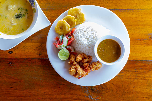 Typical traditional Ecuadorian almuerzo lunch plate presenting fried shrimp, white rice, menestra, patacones, fresh salad, a side of sauce, and a separate bowl of creamy seafood soup on a rustic wooden table