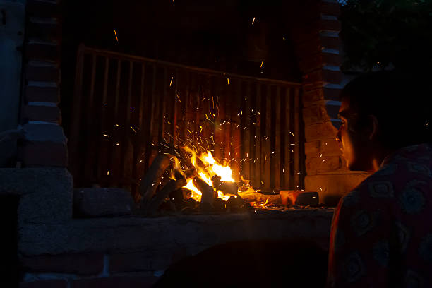 Young man watches logs burning in a rustic brick outdoor grill at night, tending embers and flames while preparing traditional South American (Ecuadorian) food for an evening gathering