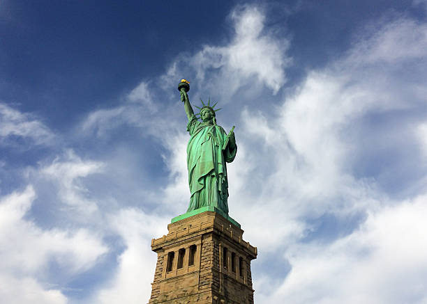A classic low-angle view of the Statue of Liberty standing prominently on its pedestal, set against a dynamic blue sky with wispy white clouds in New York City