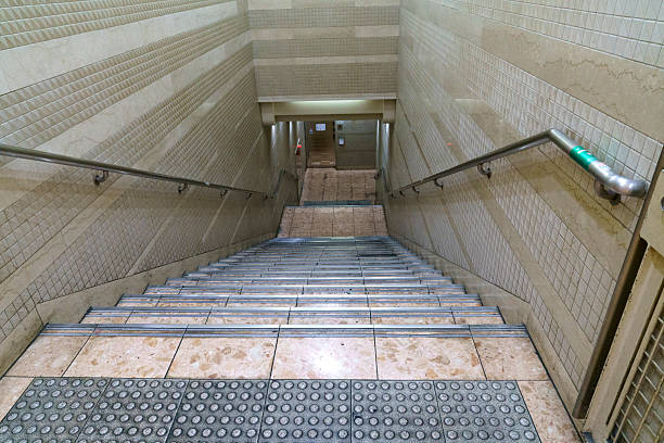 High-angle view looking down a clean tiled staircase in a modern subway station or urban underground passage. Features beige ceramic wall tiles, metal handrails, and tactile paving at the top.