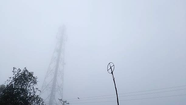 Dense fog shrouds a landscape in Punjab, highlighting the silhouette of a tall telecommunication tower. The structure, made of metal, rises visibly into the sky, intersected by power lines. A small bird perches on one of the lines. In the foreground, a tree with sparse leaves adds texture, while an antenna is visible to the right. The muted colors and obscured visibility emphasize the heavy fog.