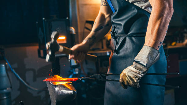 Close-up of young Caucasian male blacksmith in workshop forging heated steel on anvil with hammer, focused on crafting. Wearing protective gloves and apron, demonstrating precision and dedication in metalwork environment.